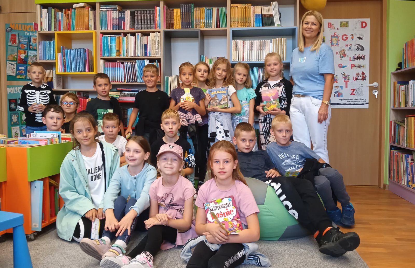 Uczniowie klasy drugiej SP w Brzezinach z wychowawczynią na tle regałów z książkami w Oddziale dla Dzieci w Bibliotece Publicznej Gminy Brzeziny
